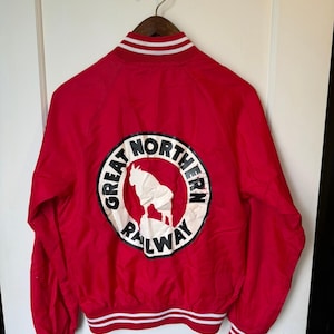 May include: A bright red bomber jacket with white striped ribbed trim on the collar, cuffs, and hem. The back features a circular white patch with a black border, displaying a white mountain goat graphic and the text Great Northern Railway.