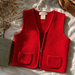 Può includere: Un gilet rosso brillante e strutturato con scollo a V e chiusura a zip completa. Il gilet presenta due tasche frontali e un'etichetta con la scritta "Stephanie Parks". L'indumento è esposto su una superficie bianca.