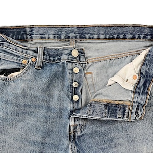 May include: Close-up of light blue denim jeans with a button fly. The jeans have a classic five-pocket design, with visible stitching and metal rivets. The fly is partially unbuttoned, revealing the interior fabric.