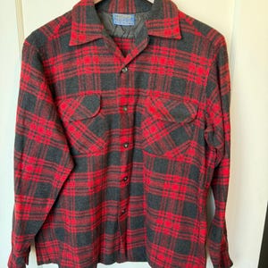 May include: A long-sleeve button-up shirt featuring a red and charcoal gray plaid pattern. The shirt has a pointed collar, two chest pockets with flaps, and a Pendleton brand label sewn into the inner neckline. It is hung on a wooden hanger.