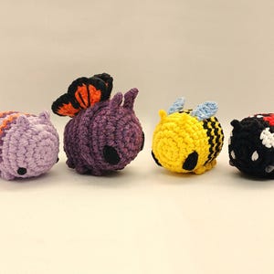 May include: Four small, round, crocheted plush toys arranged in a row. The first is light purple with orange stripes, the second is dark purple with orange and black butterfly wings, the third is yellow with black stripes, and the fourth is red and black.