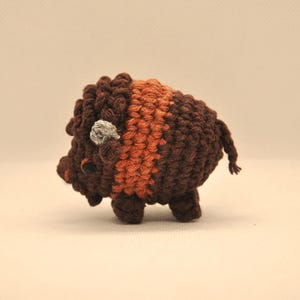 May include: A handmade crochet bison figurine. The bison is mainly dark brown with a lighter brown stripe across its back and a small grey nose. The bison has small legs and a short tail.