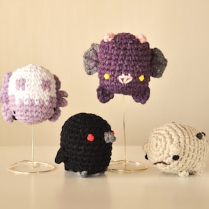 May include: Four crocheted stuffed animals on display stands. The animals are a purple axolotl, a black bird, a white axolotl, and a beige creature with black eyes.