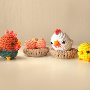 May include: A set of crocheted chickens, including a brown and green rooster, a white hen with a nest of pink eggs, and a yellow chick.