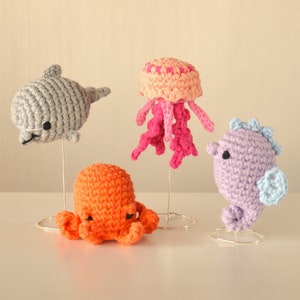 May include: Four crocheted sea creatures on wire stands: a grey dolphin, a pink jellyfish, an orange octopus, and a purple seahorse.