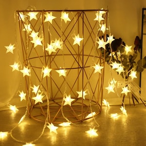 May include: A warm, inviting image of star-shaped fairy lights draped over a gold wire basket. The lights are a soft yellow, creating a cozy ambiance. The basket has a geometric design, and the lights are strung along the wire.