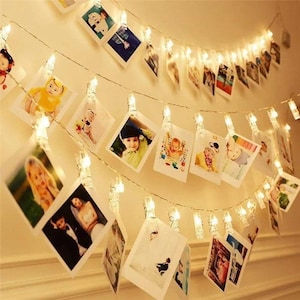 May include: A decorative string of warm white LED lights with small clear clips holding photographs. The photos show people and events. The lights are strung across a wall, creating a display.