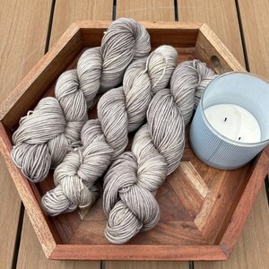 Peut inclure: Un plateau en bois hexagonal présente plusieurs écheveaux de fil gris et crème, ainsi qu'une bougie bleu clair dans un bougeoir en verre. La laine est disposée de manière attrayante, suggérant un thème artisanal ou de loisirs. La bougie ajoute une touche d'ambiance.