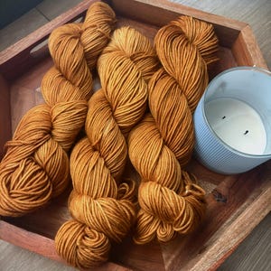 May include: A wooden tray displays several skeins of golden-brown yarn and a light blue candle. The yarn is tightly wound, and the candle has a white wax interior. The tray has a natural wood grain.