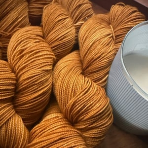 May include: A collection of golden-brown yarn skeins. The yarn is tightly wound, with visible individual strands. A partially visible container with blue stripes is in the background, suggesting a crafting theme.