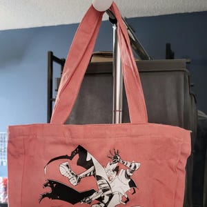 Könnte beinhalten: Eine rosa Canvas-Tragetasche mit einer Schwarz-Weiß-Illustration eines Charakters in einer dynamischen Pose. Der Charakter trägt ein weißes Hemd und eine schwarze Hose und hält eine Waffe.