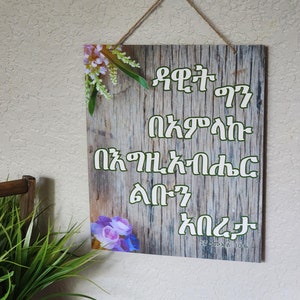 Op de afbeelding: Houten wanddecoratie met groene tekst op een houtnerf achtergrond. Bloemenaccenten in paars en wit sieren de hoeken. Een touwtje is bevestigd om op te hangen. De tekst is in een vreemde taal.