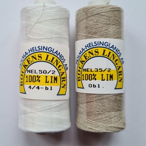 Op de afbeelding: Twee cilindrische klossen garen, een witte en een beige, elk voorzien van een etiket met de tekst Holma-Helsinglands AB Bockens Lingarn, 100% Lin. De witte klos is gelabeld NEL50/2 4/4-bl en de beige klos is gelabeld NEL35/2 0bl.