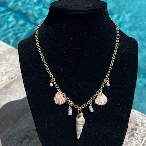 May include: A gold chain necklace with three seashell charms and white beads. The seashells are a pink and white scallop, a white scallop, and a brown and white cone shell.