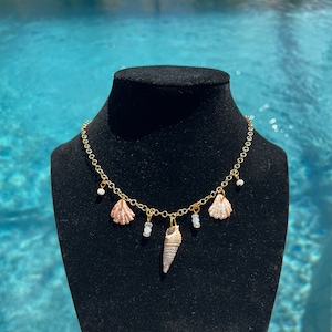May include: A gold chain necklace with three small seashells and a larger seashell pendant. The seashells are white and pink.