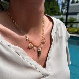 May include: A gold chain necklace with multiple shell charms, including a large seashell pendant. The necklace is worn by a person with a light brown shirt.