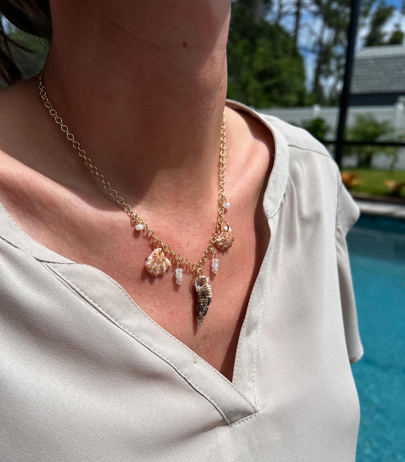 May include: A gold chain necklace with multiple shell charms, including a large seashell pendant. The necklace is worn by a person with a light brown shirt.