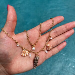 May include: A gold chain necklace with a pearl pendant and multiple shell charms. The necklace is layered with a second gold chain with small white beads.