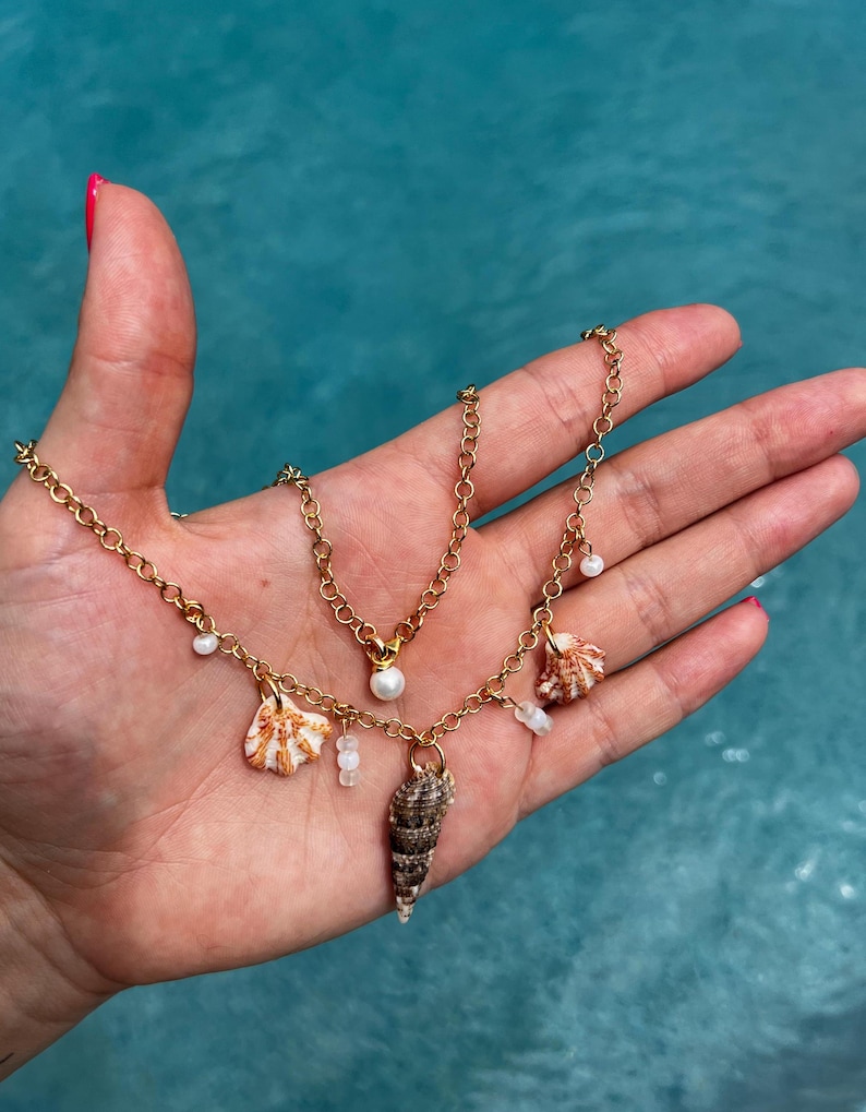 May include: A gold chain necklace with a pearl pendant and multiple shell charms. The necklace is layered with a second gold chain with small white beads.
