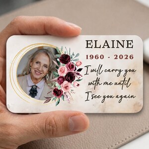 Puede incluir: Una tarjeta conmemorativa sostenida en una mano, con una foto de una mujer en un óvalo dorado. La tarjeta incluye el nombre "Elaine", las fechas 1960-2026 y el texto "I will carry you with me until I see you again."