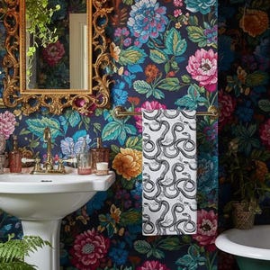 May include: A bathroom with a floral wallpaper in shades of blue, pink, and orange. A white pedestal sink with gold fixtures is in the foreground. A towel with a snake pattern hangs on a gold bar. A gold framed mirror is on the wall.
