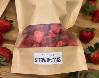 May include: A brown paper bag with a clear window, filled with freeze-dried strawberries. The label reads "Freeze-Dried STRAWBERRIES" in black text. Fresh strawberries with green leaves are scattered around the bag on a wooden surface.