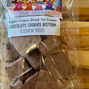 May include: A bag of vegan freeze-dried ice cream with chocolate cookies and cream flavor. The ice cream is made with a cashew base. The bag has a label with the brand name "Sugar Shockers" and a cartoon image of four girls.