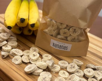 May include: A bunch of fresh yellow bananas next to a bag of freeze-dried banana slices. The bag is labeled "Bananas". Sliced bananas are scattered on a wooden cutting board, showcasing a healthy snack option.