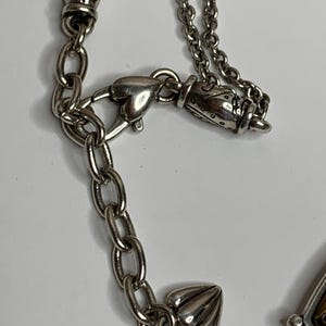 May include: Close-up of a silver necklace with detailed design elements. The necklace has a chain with a heart-shaped clasp and decorative features. It includes ornate beads and a small, detailed charm. The background is a plain white surface.