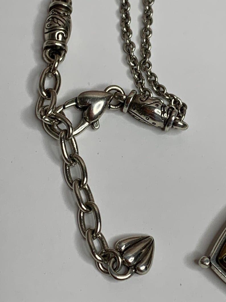 May include: Close-up of a silver necklace with detailed design elements. The necklace has a chain with a heart-shaped clasp and decorative features. It includes ornate beads and a small, detailed charm. The background is a plain white surface.