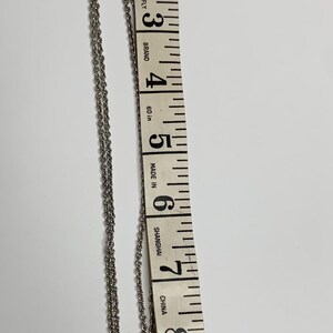 May include: Silver necklace with a square pendant, featuring an ornate design. The necklace is measured with a 25 cm measuring tape. The tape is marked with the text "Fiber-Glass", "Butterfly", "Brand", "Made in Shanghai", and "China".