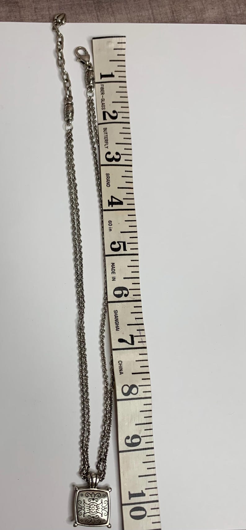May include: Silver necklace with a square pendant, featuring an ornate design. The necklace is measured with a 25 cm measuring tape. The tape is marked with the text "Fiber-Glass", "Butterfly", "Brand", "Made in Shanghai", and "China".