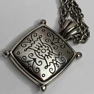 May include: Silver pendant necklace with a square, decorative pendant. The pendant has a floral design with swirling details and small, round accents. The necklace chain is silver in colour.