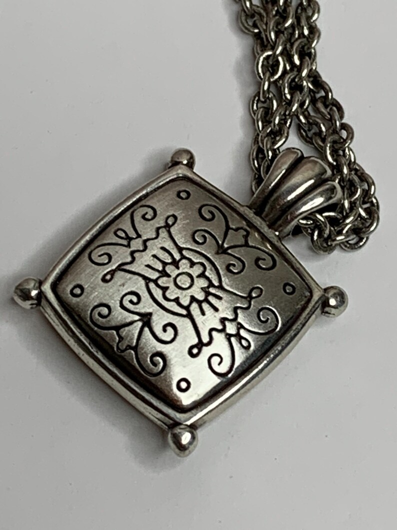 May include: Silver pendant necklace with a square, decorative pendant. The pendant has a floral design with swirling details and small, round accents. The necklace chain is silver in colour.