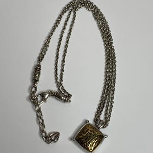 May include: Silver-tone necklace with a square pendant. The pendant has a gold-coloured centre with an ornate design. The necklace features multiple strands of chain and a lobster clasp closure.