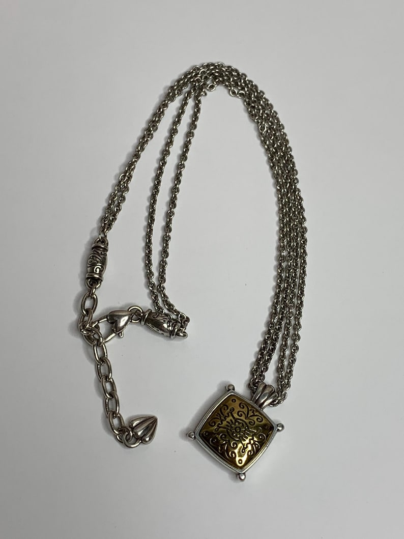 May include: Silver-tone necklace with a square pendant. The pendant has a gold-coloured centre with an ornate design. The necklace features multiple strands of chain and a lobster clasp closure.