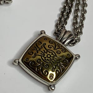 May include: Silver necklace featuring a square pendant with a gold-coloured centre and an ornate floral design. The pendant is framed by a silver-toned border. The chain is made of small, linked circles.