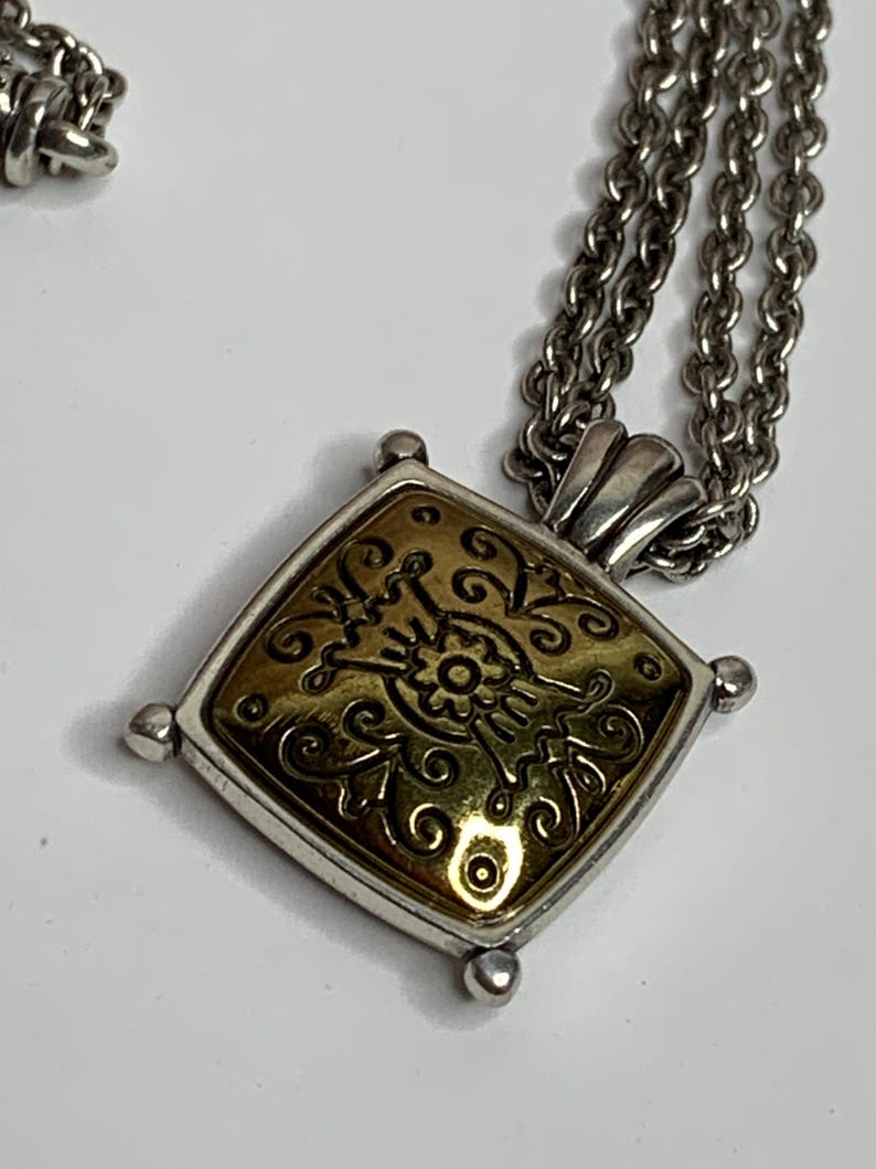 May include: Silver necklace featuring a square pendant with a gold-coloured centre and an ornate floral design. The pendant is framed by a silver-toned border. The chain is made of small, linked circles.