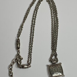 May include: Silver necklace with a square pendant, decorated with a floral pattern. The pendant is suspended from a triple-strand chain, with a decorative clasp and an adjustable chain. The necklace is shown on a white background.