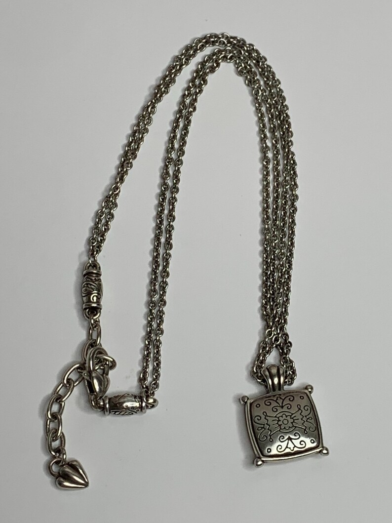 May include: Silver necklace with a square pendant, decorated with a floral pattern. The pendant is suspended from a triple-strand chain, with a decorative clasp and an adjustable chain. The necklace is shown on a white background.