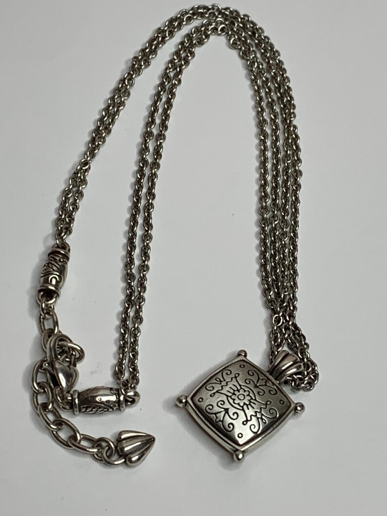 May include: Silver necklace with a square pendant, decorated with a floral pattern. The pendant hangs from a fine chain with an adjustable clasp. A classic piece of jewellery.