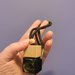 May include: A two-part car air freshener with a black and light wood cube design. The top cube is light wood, and the bottom cube is black. A black cord with a wooden bead and metal accent is attached for hanging.