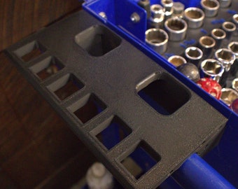 Può includere: Un organizzatore di strumenti in plastica nera con più fessure per contenere gli strumenti. L'organizzatore è attaccato a una cassetta degli attrezzi in metallo blu.