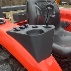 May include: A black plastic cup holder with a built-in phone mount and three switches. The cup holder is mounted on a red tractor.