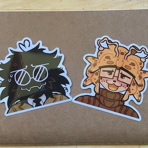 May include: Two stickers featuring cartoon characters. One has a dark green face with sunglasses and a light brown outline. The other has a light brown face with glasses and a brown turtleneck. Both stickers have a white border.