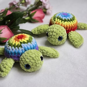 Puede incluir: Dos juguetes de peluche de tortuga hechos a crochet con caparazones de colores arcoíris. Las tortugas son verdes y tienen grandes ojos negros.