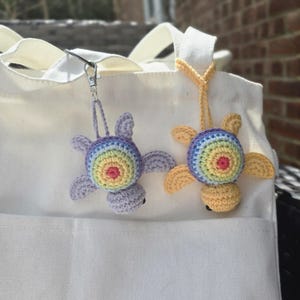 May include: Two crochet turtle keyrings. The turtles have rainbow-coloured shells and are attached to a white tote bag. One turtle is purple with a rainbow shell, and the other is yellow with a rainbow shell. The keyrings are attached to the bag with a silver clasp and a yellow string.