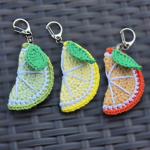 May include: Three crochet keychains shaped like citrus fruit slices: a lime, a lemon, and an orange. Each slice has a green leaf and a silver-colored clasp. The lime is green, the lemon is yellow, and the orange is orange.