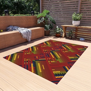 Puede incluir: Alfombra rectangular para exteriores con un patrón geométrico. La alfombra presenta una base roja con acentos de amarillo, verde y azul. El diseño incluye varias formas y líneas, creando una estética moderna. La alfombra está colocada sobre una terraza de madera.