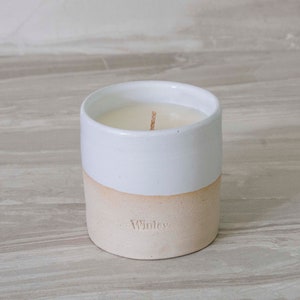May include: A white ceramic candle jar with a beige base. The jar has a single wick and the word "Winley" is printed on the base.